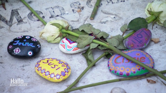 Eine Gedenktafel mit Blumen und bunt bemalten Steinen. © Screenshot 