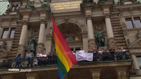 Die Regenbogenflagge, gehisst am Hamburger Rathaus, Menschen stehen auf dem Balkon und halten Plakate. © Screenshot 