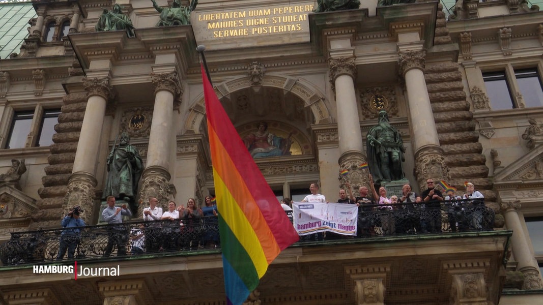 Die Regenbogenflagge, gehisst am Hamburger Rathaus, Menschen stehen auf dem Balkon und halten Plakate.