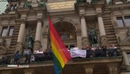 Die Regenbogenflagge, gehisst am Hamburger Rathaus, Menschen stehen auf dem Balkon und halten Plakate. © Screenshot 