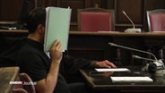 Angeklagter mit einer Mappe vor dem Gesicht im Gerichtssaal. © Screenshot 