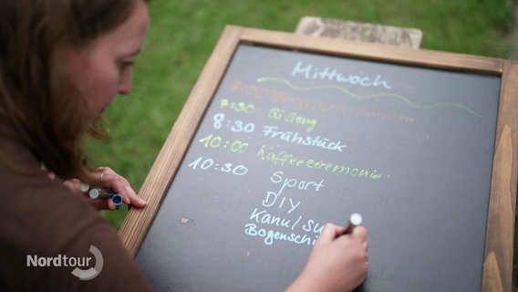 Eine Camp-Mitarbeiterin schreibt den Tagesablaufen und angebotene Aktivitäten auf eine Kreidetafel. Auf dem Proigramm stehen unter anderem Sport, diy, Kanufahren und Bogenschießen. © Screenshot 