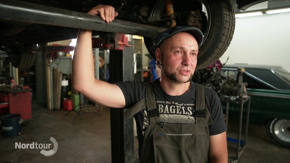Schrauber Jan Sailer lehnt in seiner Werkstatt an einem empor gehobenem Wagen. Er trägt Latzhose und Schiebermütze. © Screenshot 