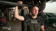 Schrauber Jan Sailer lehnt in seiner Werkstatt an einem empor gehobenem Wagen. Er trägt Latzhose und Schiebermütze. © Screenshot 