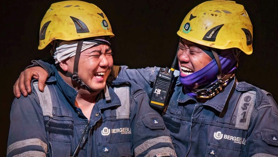 Zwei Männer in Arbeitsmontur und gelben Helmen liegen sich lachend in den Armen. © Screenshot 