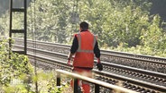 Ein Mann in orange-farbener Sicherheitskleidung läuft an Bahngleisen entlang. © Screenshot 