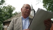 Ein älterer Herr, Vorstand einer katholischen Gemeinde, steht vor einer Kirche und betrachtet Unterlagen, die er in den Händen hält. © Screenshot 