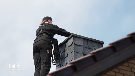 Eine Person steht in schwarzer Arbeitskleidung auf einem Dach an einem Schornstein. © Screenshot 