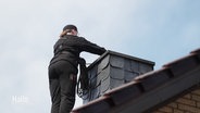 Eine Person steht in schwarzer Arbeitskleidung auf einem Dach an einem Schornstein. © Screenshot 