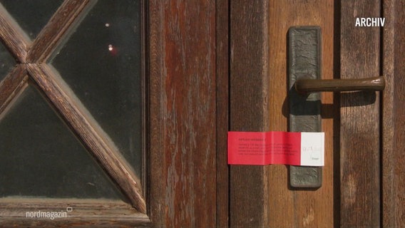 Auf dem Schloss einer Holztür ist ein rot-weißer behördlicher Aufkleber angebracht. © Screenshot 