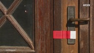 Auf dem Schloss einer Holztür ist ein rot-weißer behördlicher Aufkleber angebracht. © Screenshot 