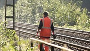 Ein Mann in orange-farbener Sicherheitskleidung geht an Bahngleisen entlang. © Screenshot 