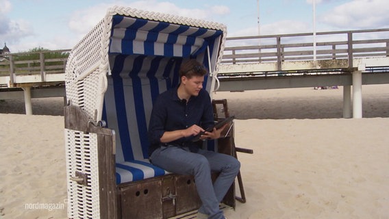 Ein junger Mann sitzt in einem Strandkorb und schaut auf ein Tablet. © Screenshot 