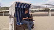 Ein junger Mann sitzt in einem Strandkorb und schaut auf ein Tablet. © Screenshot 