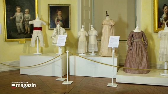 Historische Kinderkleider in einer Ausstellung. © Screenshot 