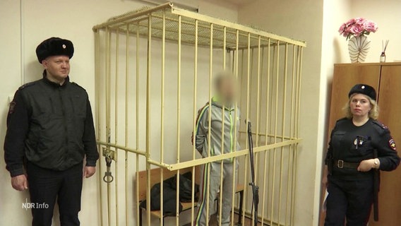 Ein Mann steht in einem Käfig, sein Gesicht ist verpixelt, neben ihm Wachleute. © Screenshot 