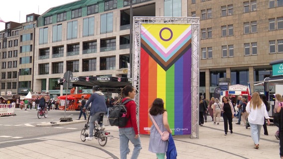 Menschen gehen während des CSD-Straßenfests in Hamburg an einem Banner in Regenbogenfarben vorbei. © Screenshot 