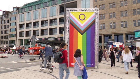 Menschen laufen beim CSD-Straßenfest in Hamburg an einem Banner in Regenbogenfarben vorbei. © Screenshot 