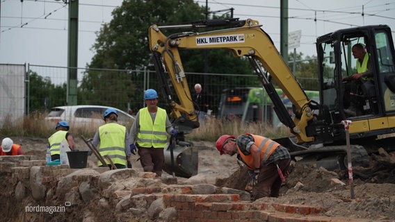 Mehrere Menschen und ein Bagger sind an Ausgrabungen einer Mauer beteiligt. © Screenshot 