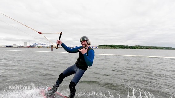 Wakeboarder Janko Borgward steht auf seinem Board und lächelt in die Kamera, während er durchs Wasser gezogen wird. © Screenshot 