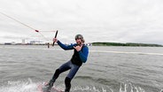 Wakeboarder Janko Borgward steht auf seinem Board und lächelt in die Kamera, während er durchs Wasser gezogen wird. © Screenshot 