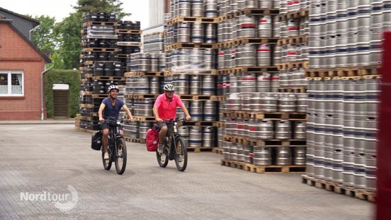 Zwei Radfahrer fahren an aufgestapelten Paletten mit Bierfässern vorbei. © Screenshot 