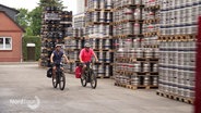 Zwei Radfahrer fahren an aufgestapelten Paletten mit Bierfässern vorbei. © Screenshot 