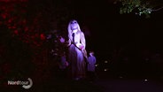 Eine große, von mehreren Performern geführte Puppe in Gestalt einer jungen Frau in historischer Kleidung, wird im Dunkeln dramatischen  angeleuchtet. © Screenshot 