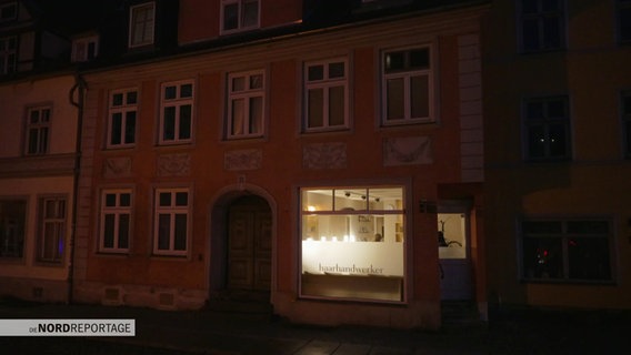 Ein Haus bei Nacht, alles ist dunkel, nur hinter einem Schaufesnter mit der Aufschrift "Haarhandwerker" brennt Licht. © Screenshot 