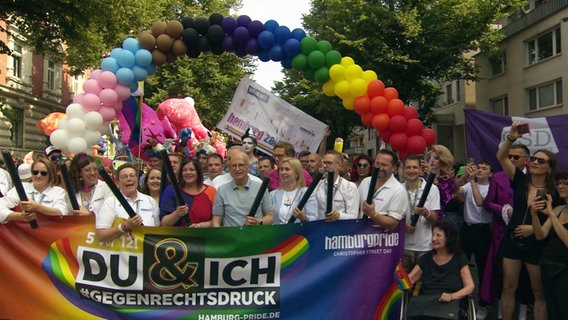 Menschen unter einem Regenbogen aus Luftballons und vor einem Plakat. © Screenshot 