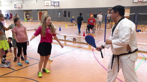 In einer Sporthalle probieren Kinder und Jugendliche mit unterschiedlichen körperlichen Behinderungen verschiedene Sportarten aus. Ein Taekwondo-Trainer hält ein Kick-Paddel, ein junges Mädchen macht sich bereit, dagegen zu treten. © Screenshot 