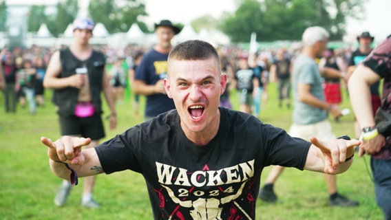 Ein junger Mann mit Wacken-T-Shirt zeigt das Wacken-Zeichen in die Kamera. © Screenshot 