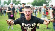 Ein junger Mann mit Wacken-T-Shirt zeigt das Wacken-Zeichen in die Kamera. © Screenshot 