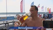 Ein Müritz-Schwimmer in Badehose trinkt nach seinem sportlichen Einsatz aus einem riesigen Bierglas. © Screenshot 
