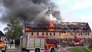 Die Feuerwehr ist mit Löschfahrzeugen vor dem brennenden Haus im Einsatz. Aus Fenstern und Dachstufl schlagen Flammen und eine große Rauchwolke steigt auf. © Screenshot 