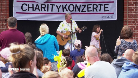Ein Kinderliedermacher und ein kleines Mädchen stehen auf einer Bühne und singen bei einem Charity-Konzert. © Screenshot 