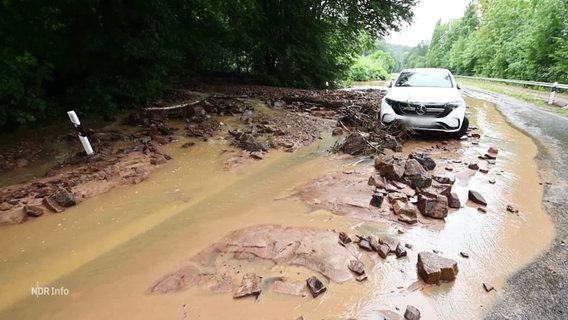 Wasser, Schlamm und Steine blockieren die Straße vor einem stehenden Auto. © Screenshot 