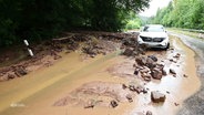 Wasser, Schlamm und Steine blockieren die Straße vor einem stehenden Auto. © Screenshot 