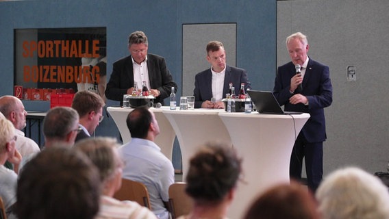 Drei Männer stehen an Rednerpulten vor einer kleineren Menge sitzender Menschen in einer Sporthalle in Boizenburg. mit dabei v.L.n.R. der Wirtschaftsminister von Mecklenburg-Vorpommern, Reinhard Meyer (SPD), und Alexander Kaczmarek, der Konzernbevollmächtigte der Deutschen Bahn für unter anderem Mecklenburg-Vorpommern. © Screenshot 