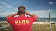 Eine Rettungskraft der DLRG hält am Strand nach Menschen in Not Ausschau. © Screenshot 