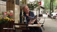 Daniel Plettenberg sitz an einem Tisch und liest ein Buch. © Screenshot 