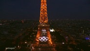 Der orange beleuchtete Eiffelturm mit dem Olympia Zeichen. © Screenshot 