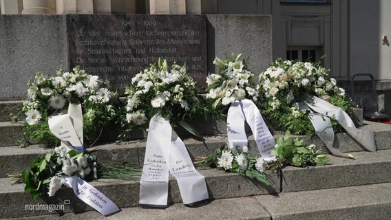 Vier grüne Kränze mit weißen Blumen liegen vor der Gedenkstätte. © Screenshot 