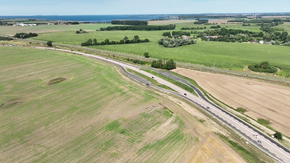 Ein Drohnenfoto eines Parkplatzes auf Rügen. Zu sehen ist eine asphaltierte Schleife, die sich beidseitig an eine Bundesstraße inmitten trockenen Graslands anschließt. Auf der Straße ist nicht wenig Verkehr. Im Hintergrund sind Felder, Gehöfte und einige Baumgruppen zu sehen. Noch weiter dahinter das Meer. © Screenshot 