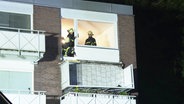 Blick auf die Fassade eines Mehrfamilienhauses. Der Fokus liegt auf einer Wohnung, wo der Balkon abgesackt nach unten hängt. In der beleuchteten Wohnung stehen Feuerwehrleute. © Screenshot 