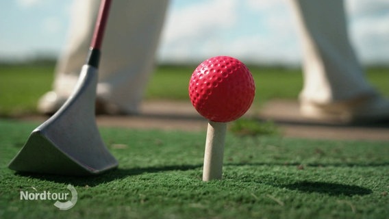 Nahaufnahme: Eine Person steht mit breitem Stand vor einem Golfball der auf dem "Tee" positioniert ist. Daneben ist ein SwinGolf-Schläger abgestellt. © Screenshot 