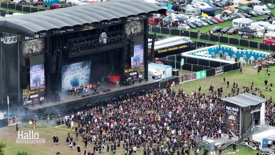 Blick aus der Vogelperspektive auf eine Menschenmenge vor einer größeren Open-Air-Bühne auf einem Hard-Rock-Festival. © Screenshot 