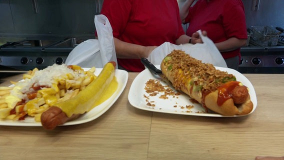 Eine Currywurst mit Pommes und ein Hotdog liegen auf einer Theke. © Screenshot 