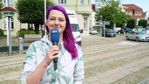 Moderatorin Dania Behm steht mit einem Mikrofon auf einem Marktplatz und grinst in die Kamera. © Screenshot 