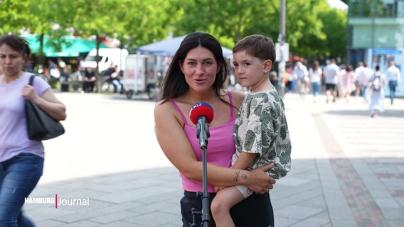 Eine junge Frau mit einem kleinen Jungen auf dem Arm steht an einem Mikrofon. © Screenshot 